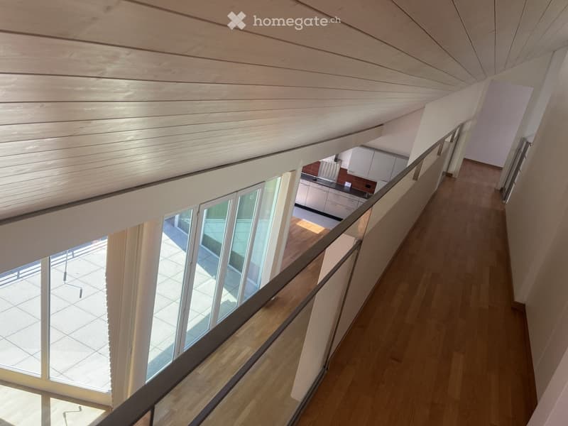 Spacious hallway with wooden flooring, modern railing, and large windows overlooking a tiled outdoor area. The ceiling is slanted with wooden paneling, and a small kitchen area is visible in the background.