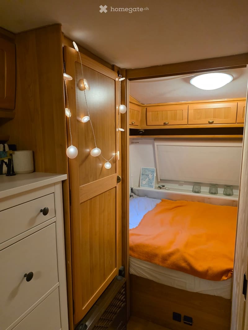 The image shows a compact bedroom with a bed, a cabinet, and a picture on the wall. The room is well-lit with a ceiling light and string lights hanging from the door.