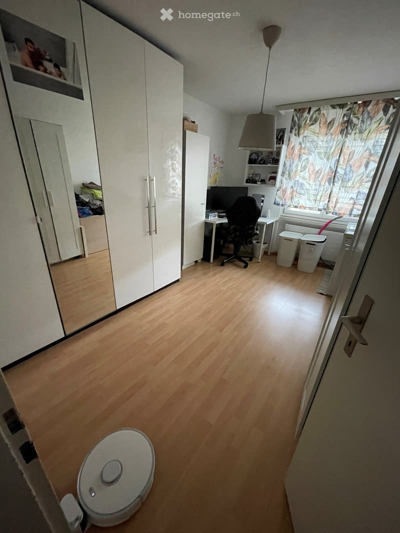 A well-lit room with wooden flooring, a large wardrobe, a desk with a computer, a chair, and a lamp hanging from the ceiling.