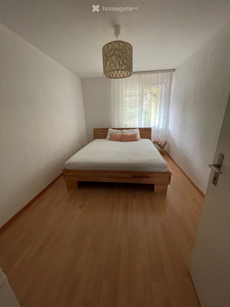 A well-lit bedroom with a wooden bed, white bedding, pink pillows, a small flower vase, and a hanging light fixture.