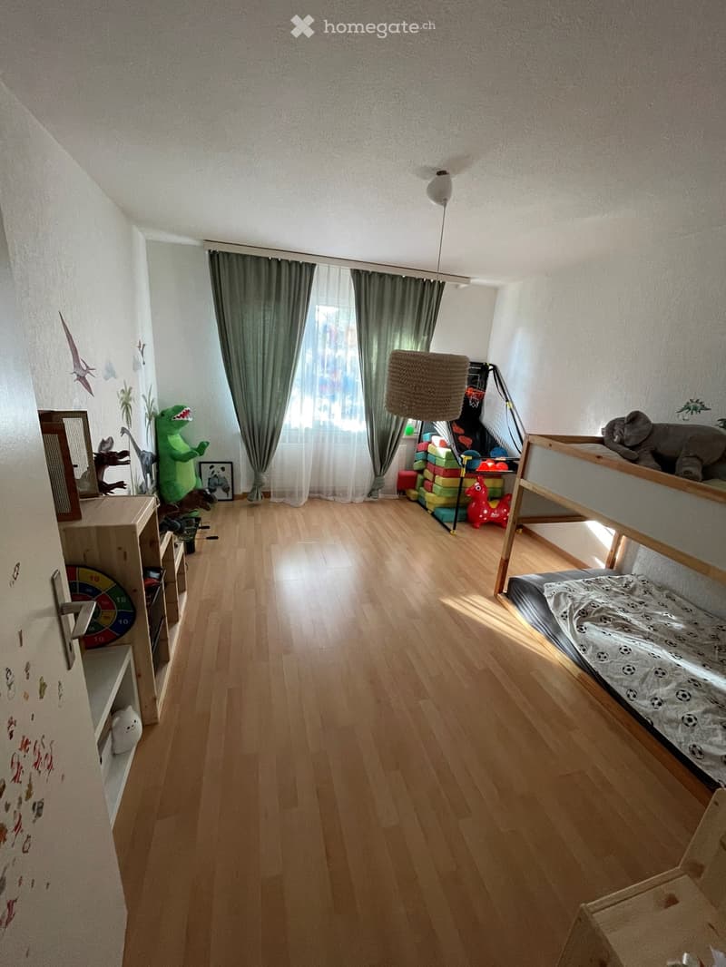 Bright, child-friendly bedroom with wooden flooring, large window with curtains, bunk bed, cat tree, and various toys.