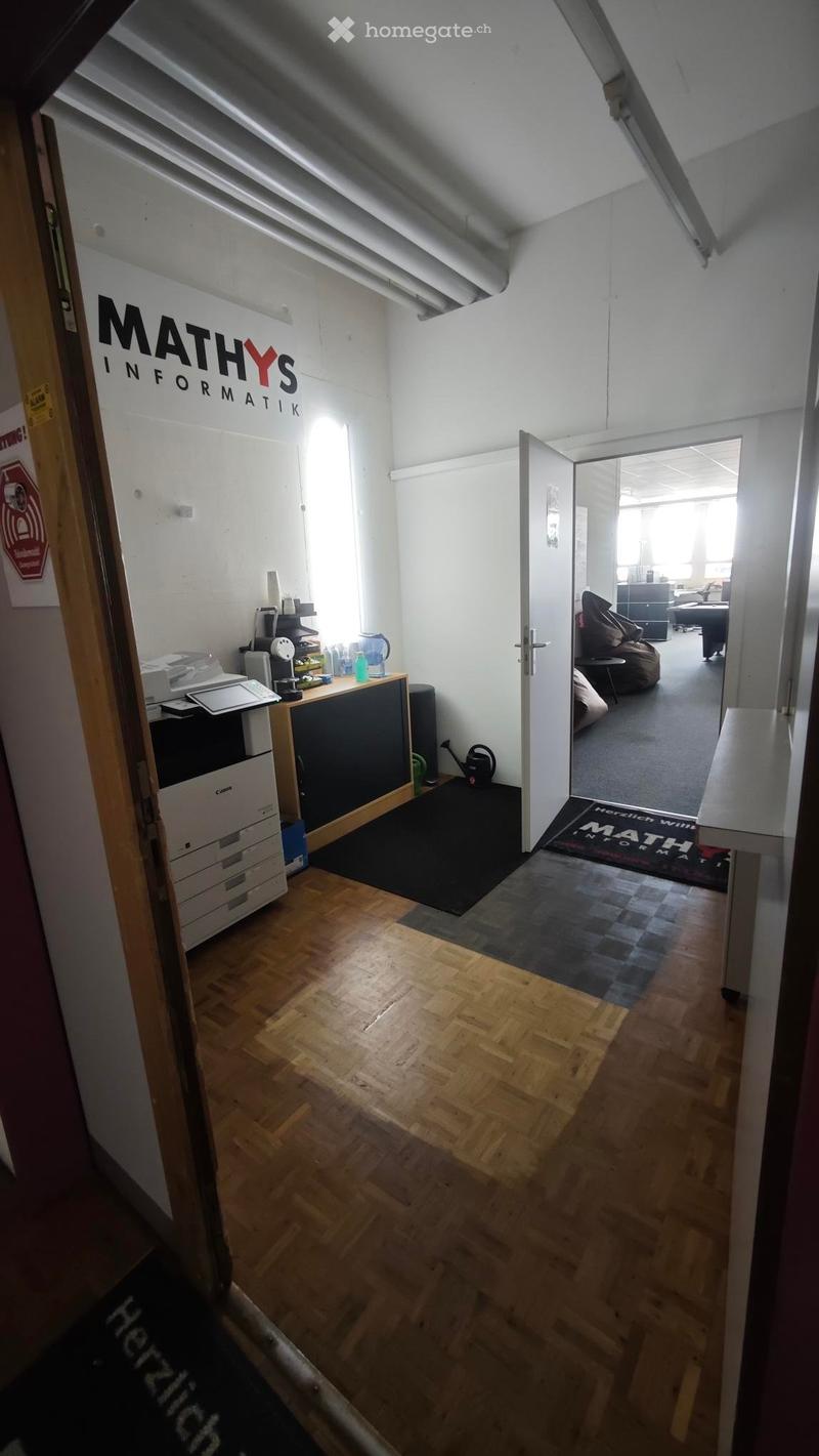 Office space with wooden flooring, a reception area with a printer and table, and a lounge area with a sofa and stool.