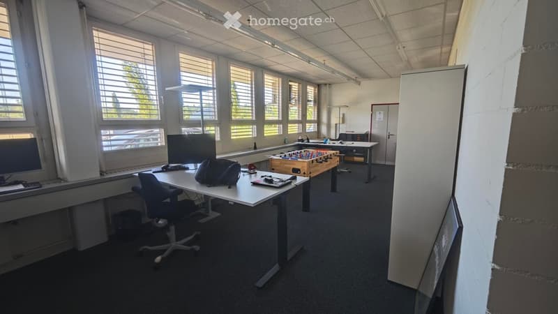 Office space with large windows, desks, chairs, a screen, a keyboard, a mouse, and a small football table.