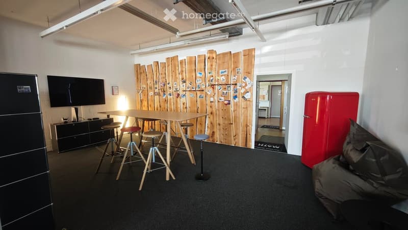 Modern, furnished room with a wooden table, stools, a red refrigerator, a large screen, and a comfortable sofa.