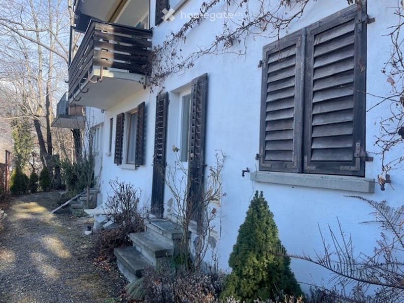 Two-story white building with wooden shutters, a balcony on the upper floor, and a small garden area with steps leading up to the entrance.