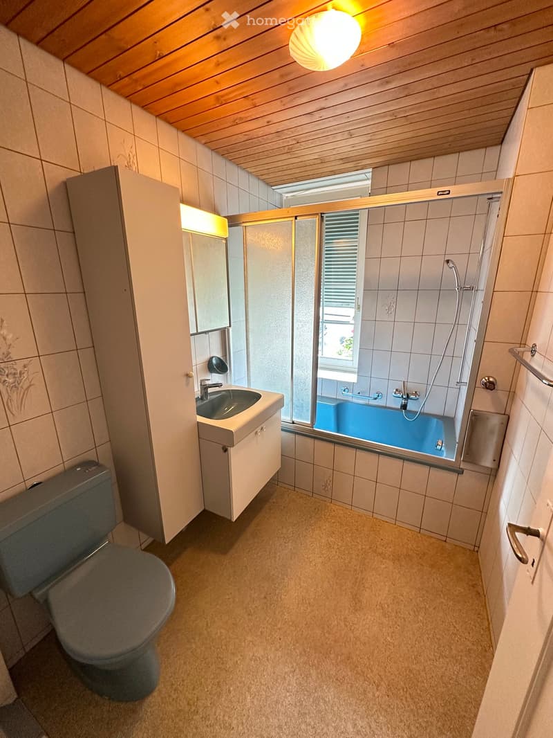 Bathroom with a toilet, sink, bathtub, and a mirror. The room features tiled walls and a wooden ceiling.