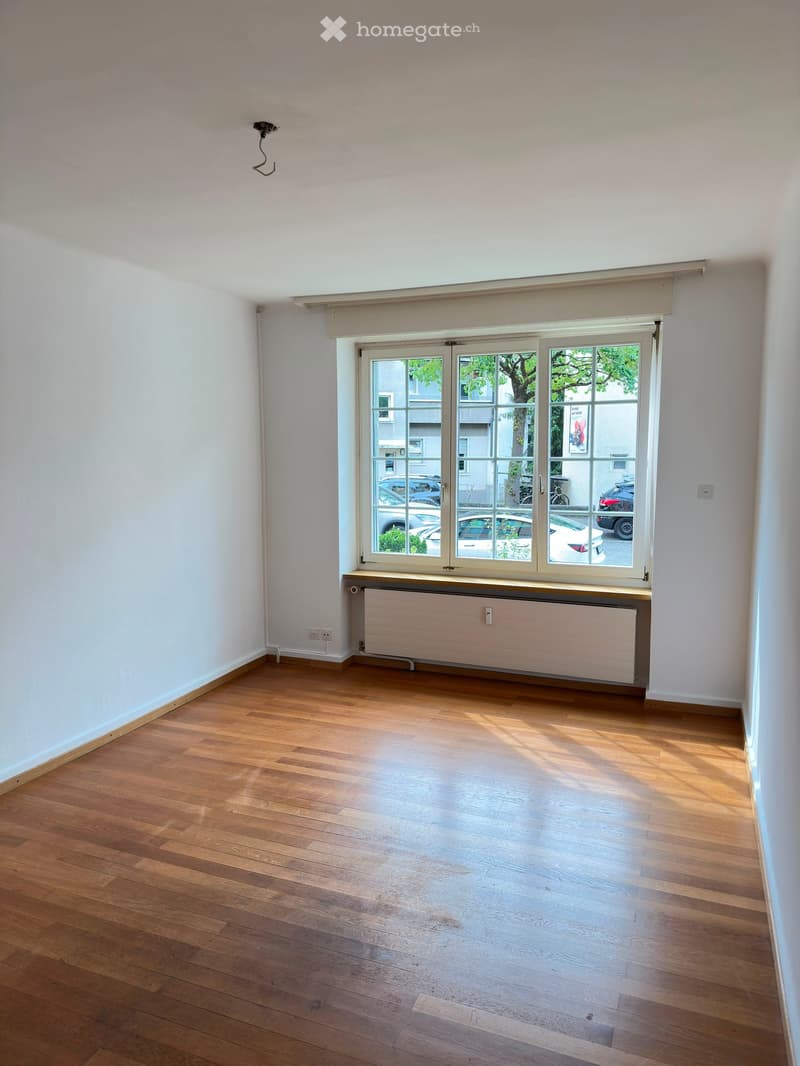 Empty room with wooden flooring, white walls, large window with blinds, radiator below the window, and visible electrical outlets.