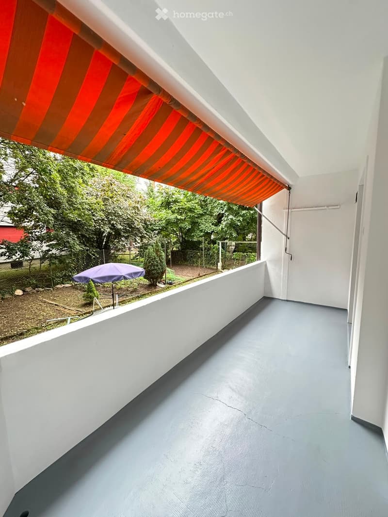 Balcony with grey floor, white walls, and an orange and brown striped awning. A purple umbrella is visible in the background garden area.