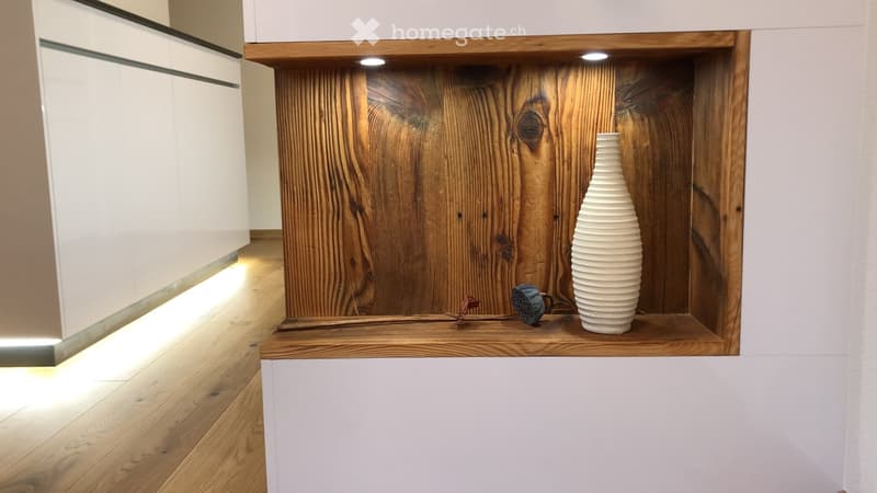 A wooden shelf with a textured wooden back wall, displaying a white vase and a blue mug, illuminated by integrated lighting.