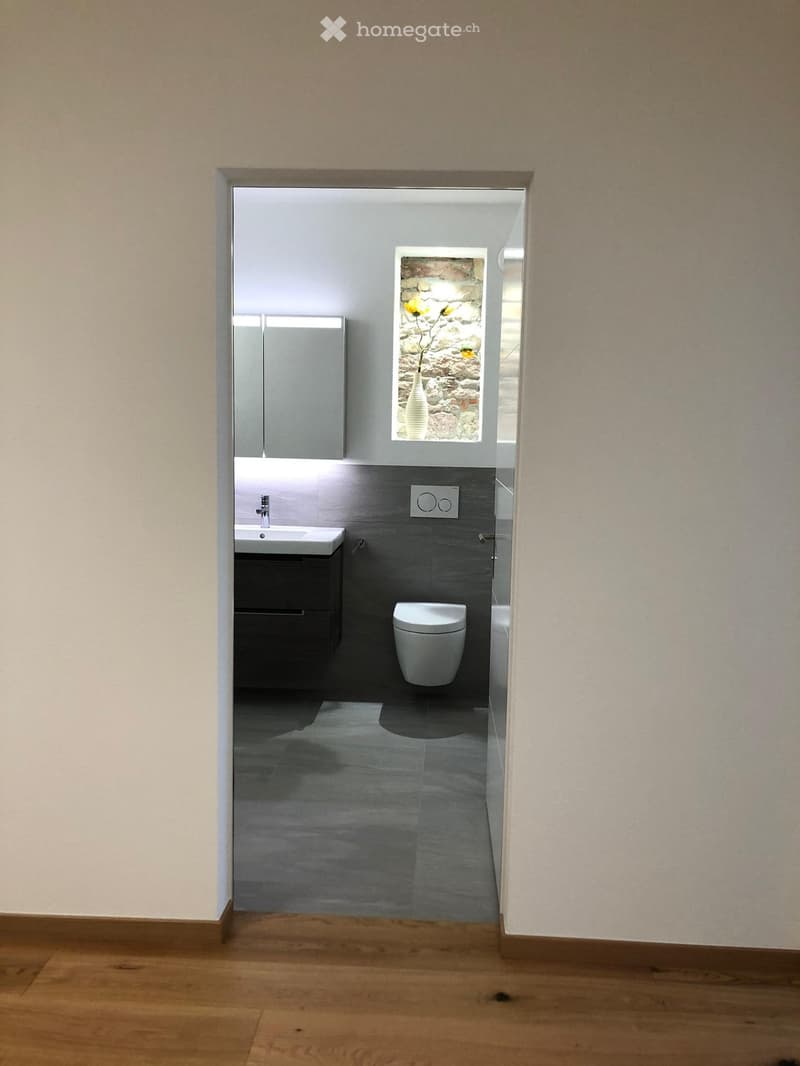 Modern bathroom with a white sink, dark cabinet, toilet, and a vase with yellow flowers on a window sill.