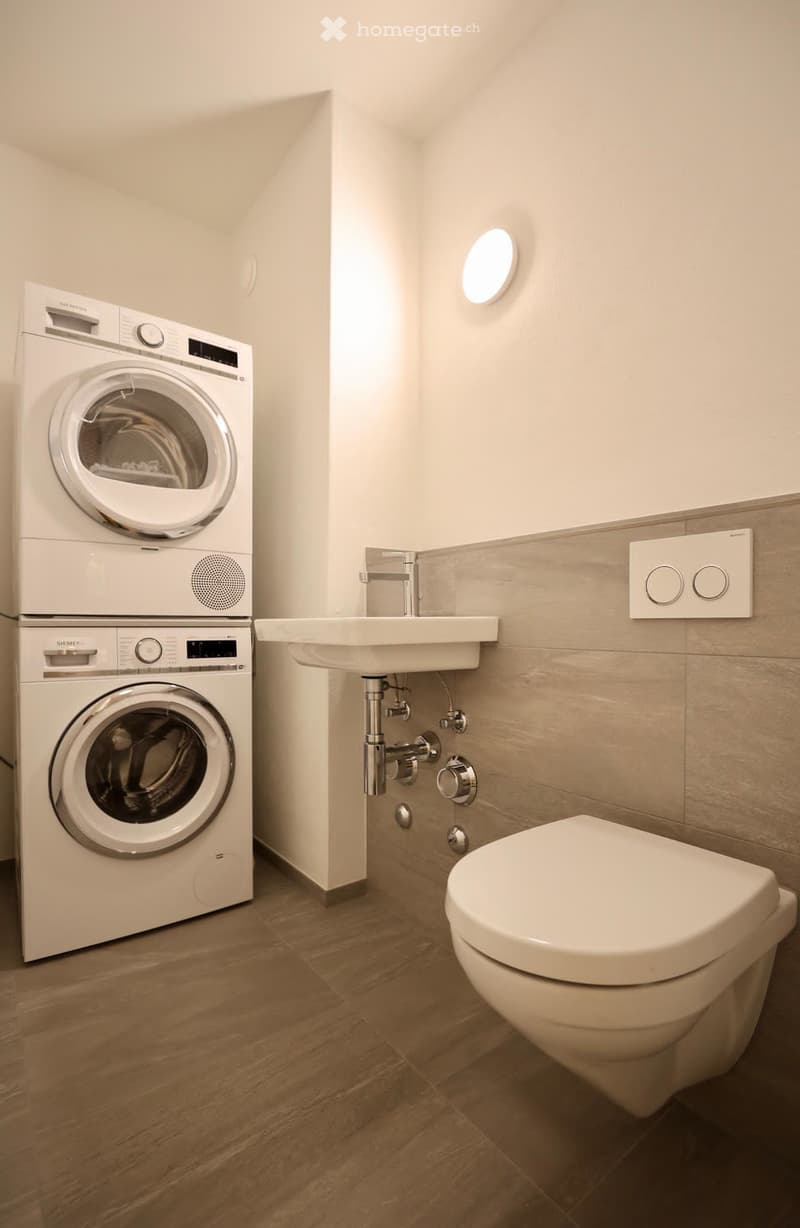 Modern bathroom with wooden flooring, white washing machine and dryer, sink with chrome faucet, and toilet.