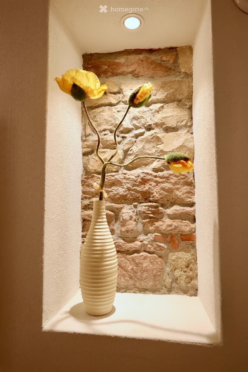 A small, well-lit alcove with a textured stone wall backdrop, featuring a white ceramic vase holding yellow flowers.