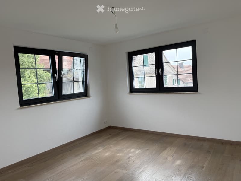 Empty room with light wooden flooring, white walls, and two large windows with black frames. Natural light streams in, illuminating the space.