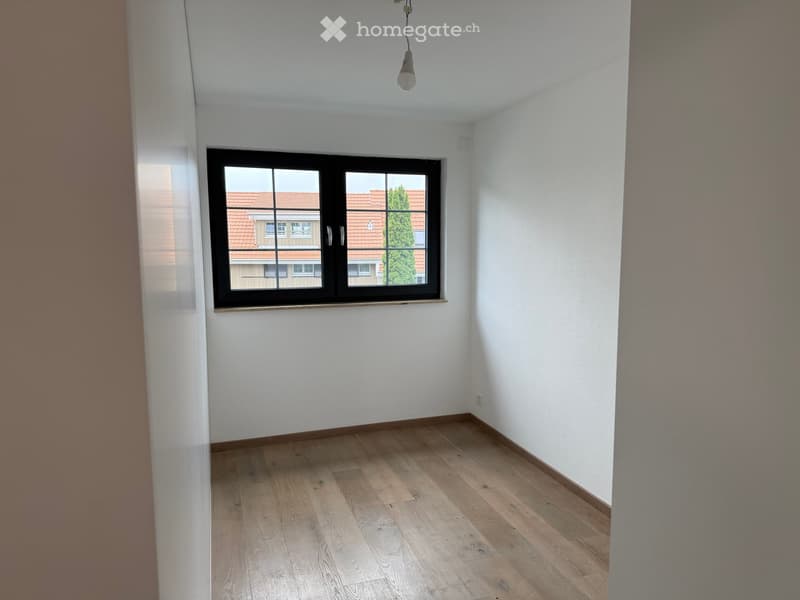 Empty room with wooden flooring, white walls, and a single window showing a view of neighboring buildings and a tree.
