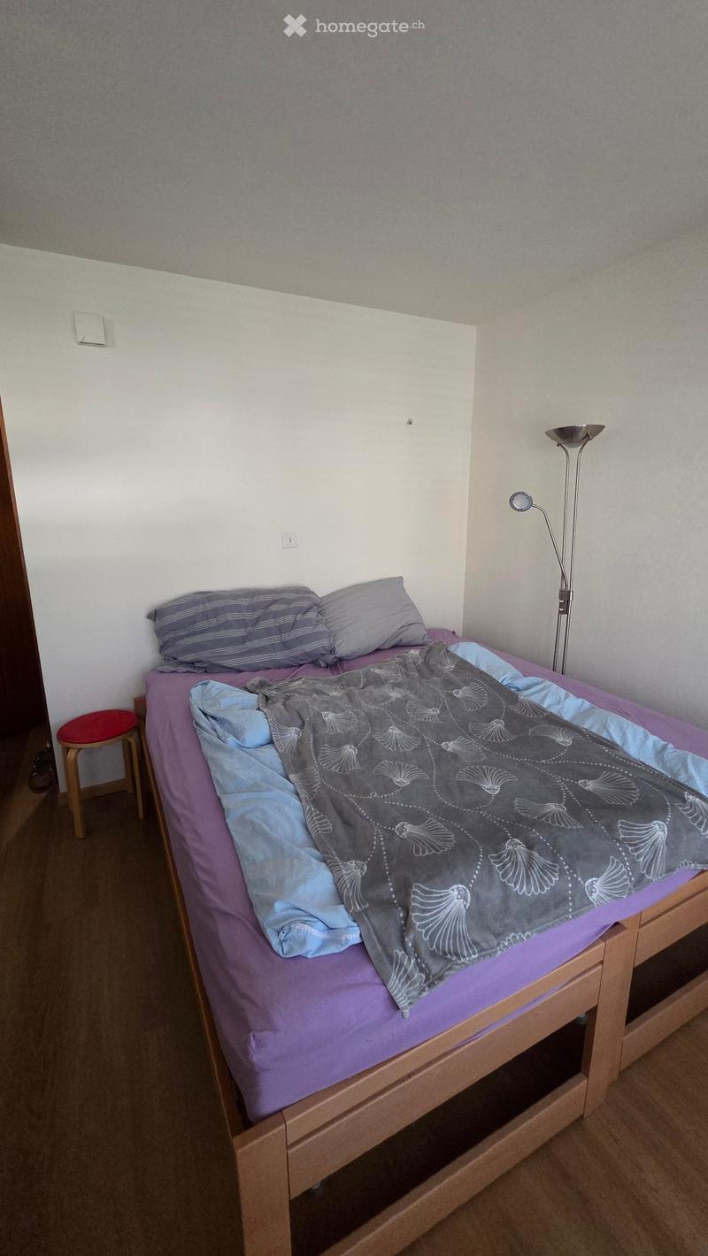 A small bedroom with a single bed, pillows, a blanket, a wooden stool, and a floor lamp. The walls are painted in a light grey color, and the floor is covered with a dark brown carpet.