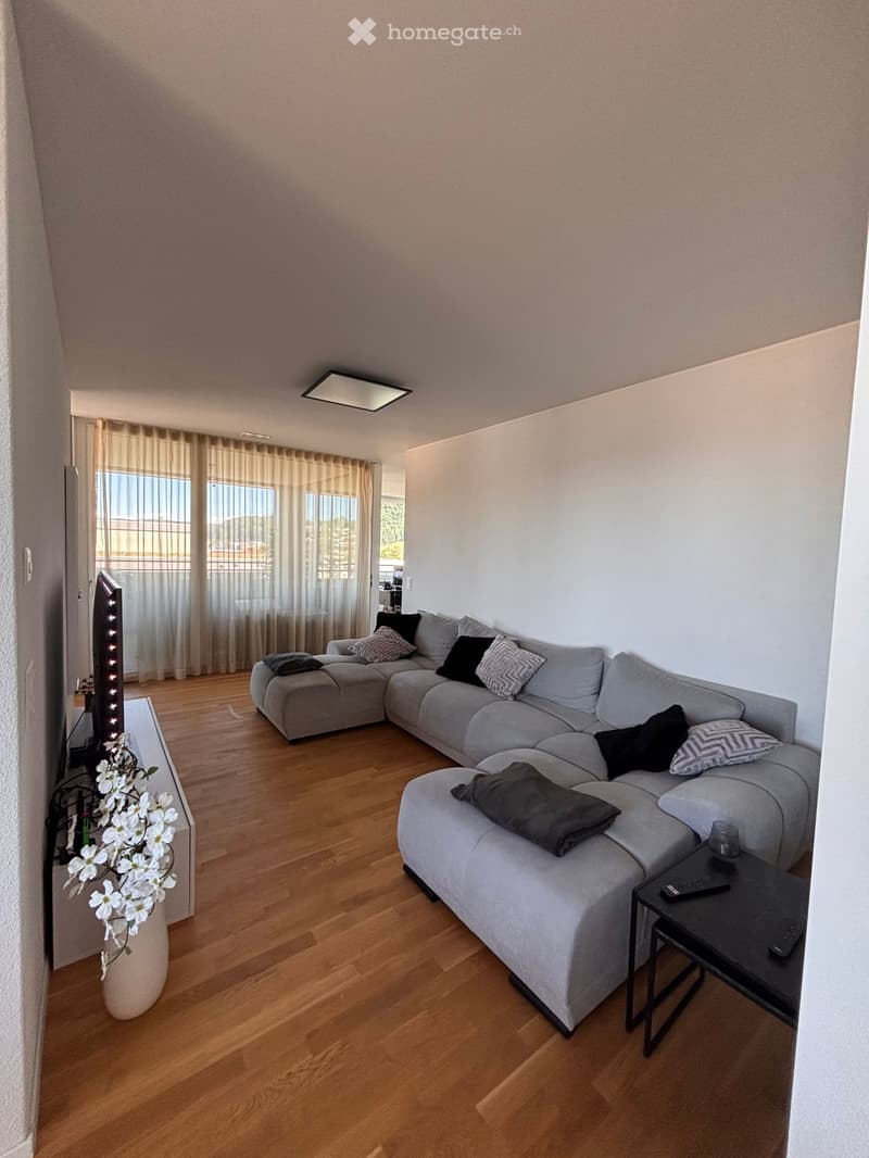 Modern living room with a grey sectional sofa, wooden flooring, large windows with sheer curtains, and a small black side table with a glass top.