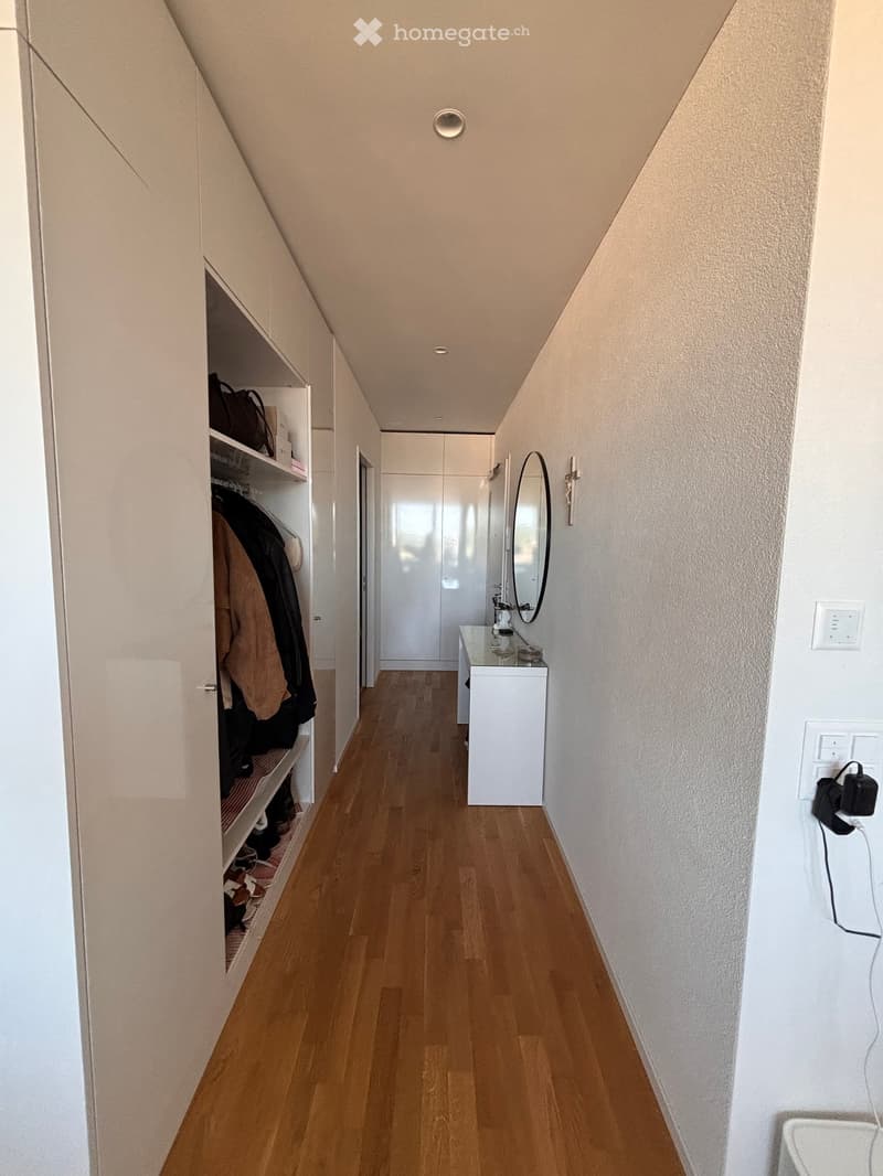 A narrow hallway with wooden flooring, white walls, built-in closet with hanging clothes and shoes, and a small sink area with a mirror and countertop.