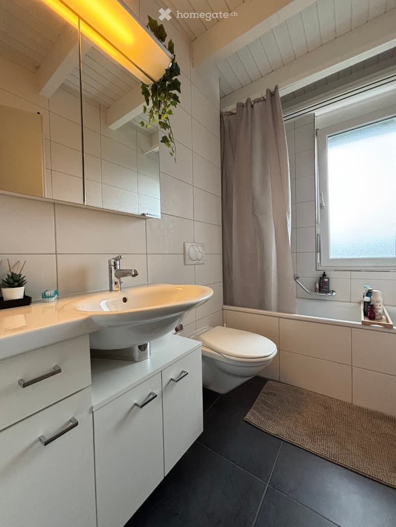 Modern bathroom with white tiles, a large sink with a modern faucet, a toilet, a bathtub with a shower curtain, and a window providing natural light. The space includes a cabinet with drawers, a mirror, and a small plant on the counter.