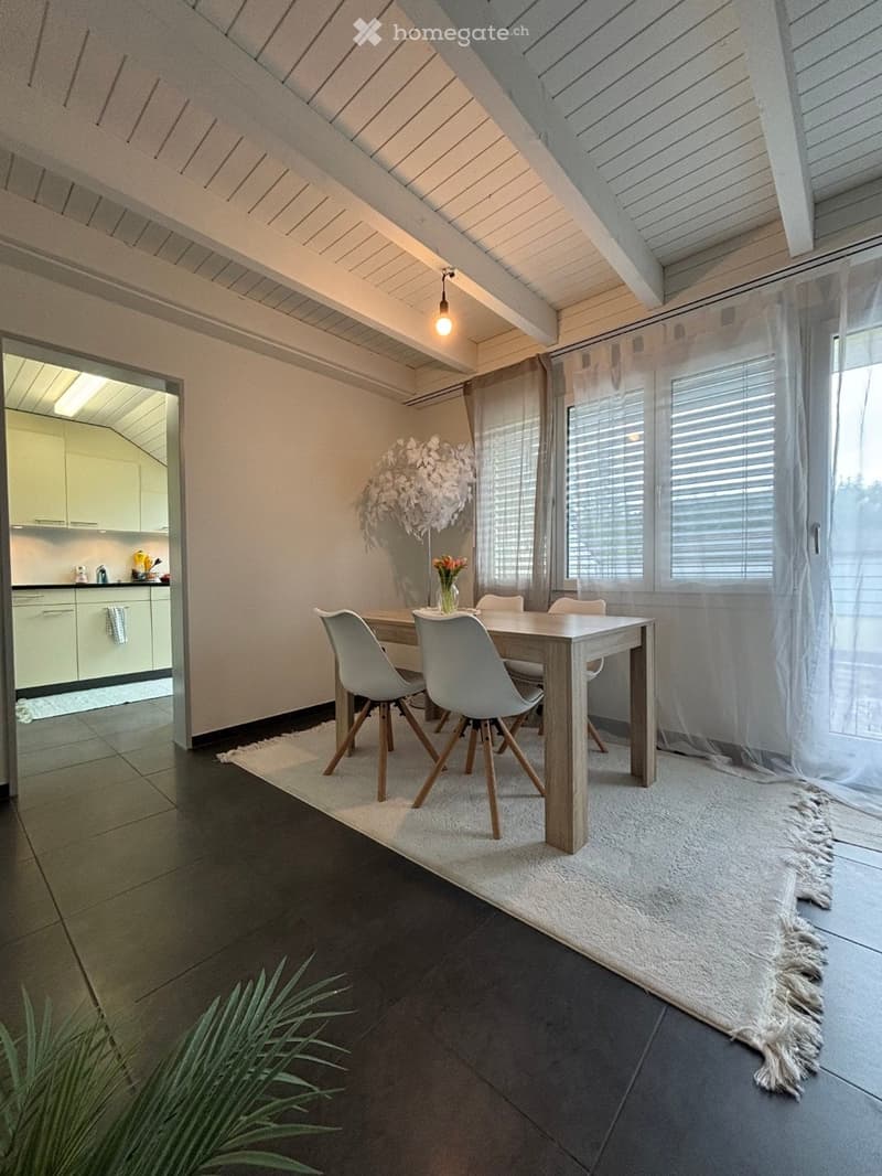 Modern dining area with a wooden dining table, four grey chairs, a white rug, and a kitchen area visible in the background.