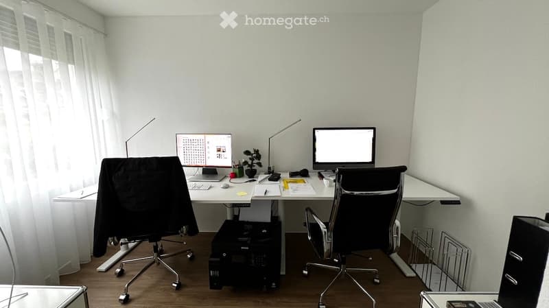 A modern, well-lit office space with a large desk, two chairs, dual monitors, a printer, and various office supplies.