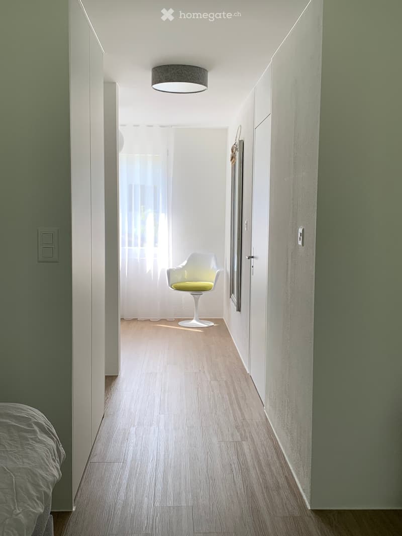 Modern hallway with light wooden flooring, white walls, and a single hanging light fixture. A single chair is placed near a doorway leading to another room.