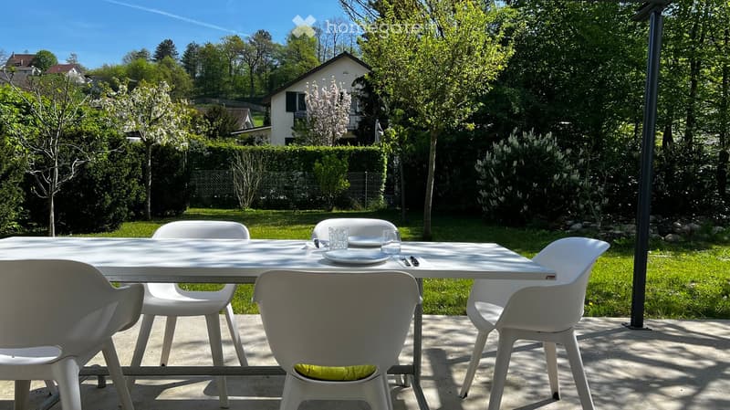 A sunny outdoor dining area with a white table and four white chairs, set on a concrete patio surrounded by a well-maintained lawn and trees.