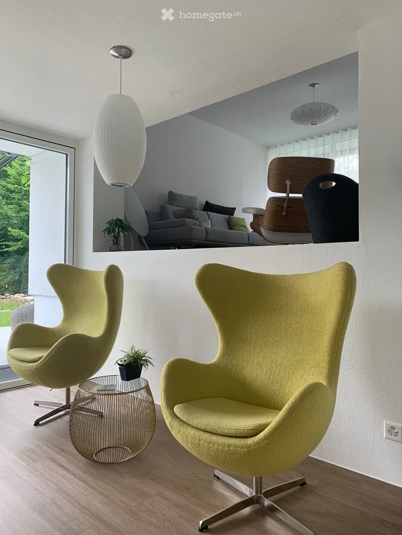 Modern living room with two green egg-shaped chairs, a small round metal table, a potted plant, and a large pendant light. A mirror reflects a grey sofa with multiple cushions and additional seating.