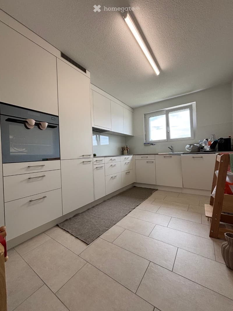 Modern kitchen with white cabinets, tiled floor, and a window above the sink. Features an oven, sink, and various kitchen items on the counter.