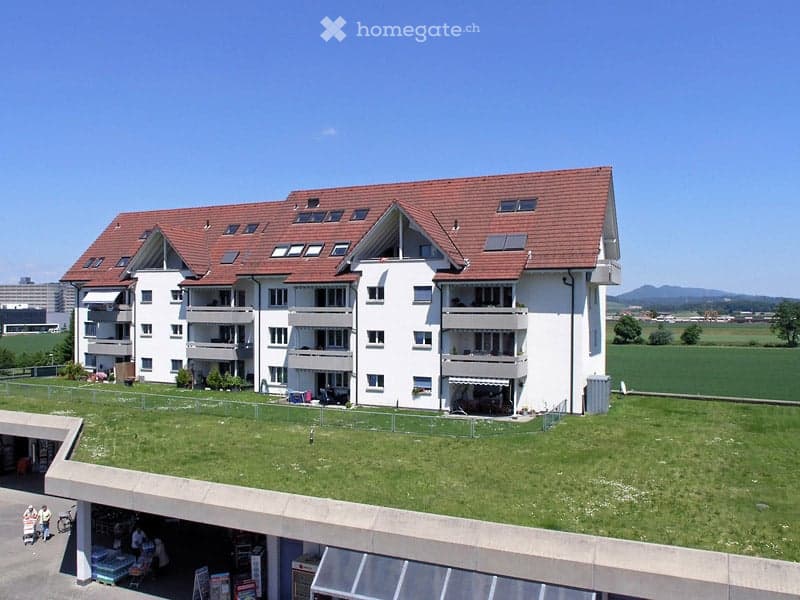 Multi-story residential building with white exterior, red-tiled roof, multiple balconies, and a green rooftop area.