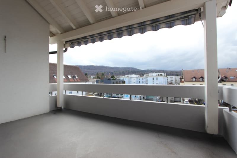 Balcony with a covered area, white railings, and a view of a town with buildings and hills in the background.