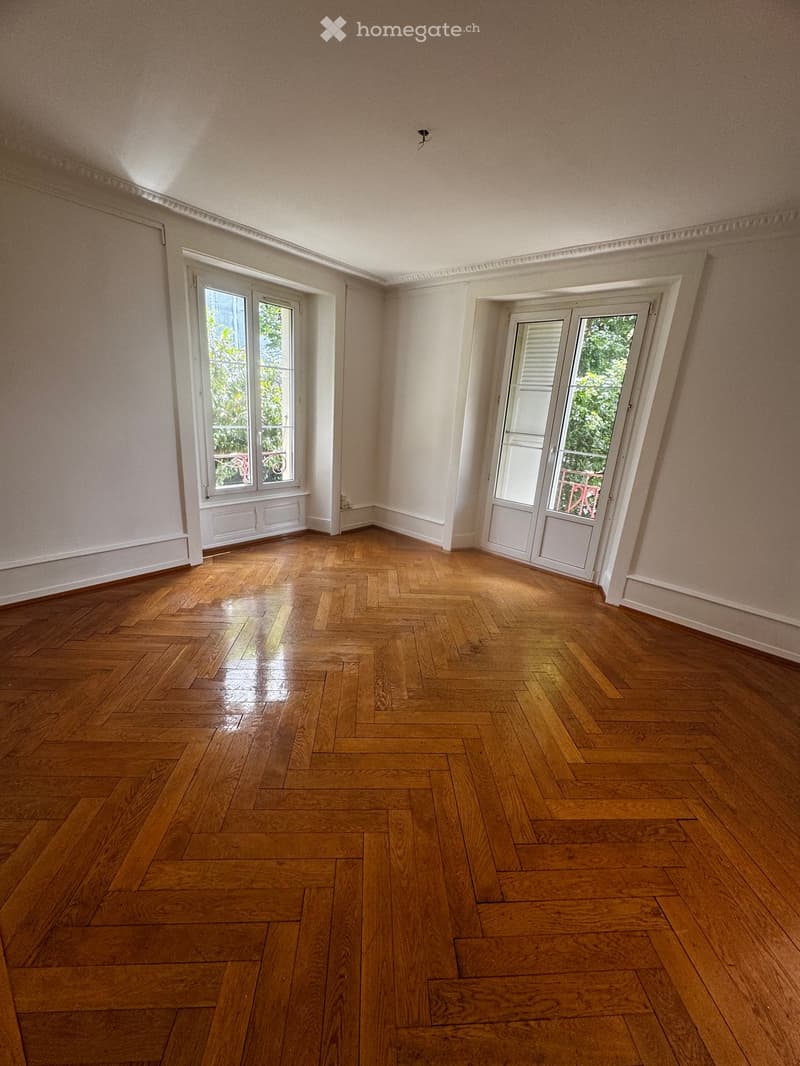 Empty room with wooden herringbone flooring, white walls, and large windows with white frames. The room has a door leading to a balcony or terrace area.