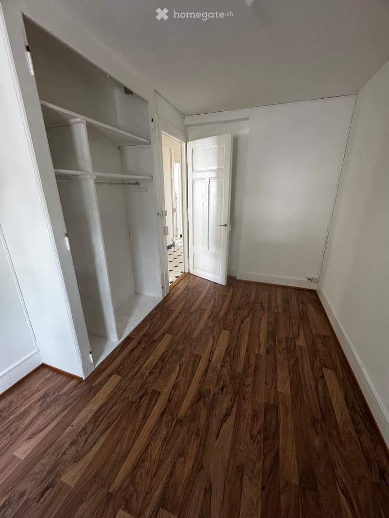Empty room with wooden flooring, white walls, and an open closet with empty shelves. A white door is visible leading to another room.