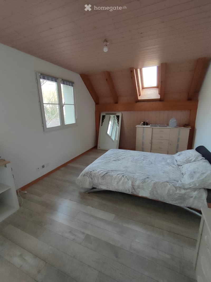 Bedroom with wooden slat flooring, slanted wooden ceiling, large windows, bed with patterned bedding, mirror, and cabinet with drawers.