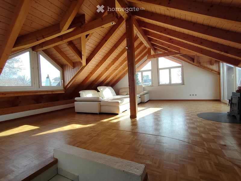 Bright, airy attic space with wooden beams and exposed ceiling, featuring a white sectional sofa and ottoman, large windows, and polished wooden flooring.
