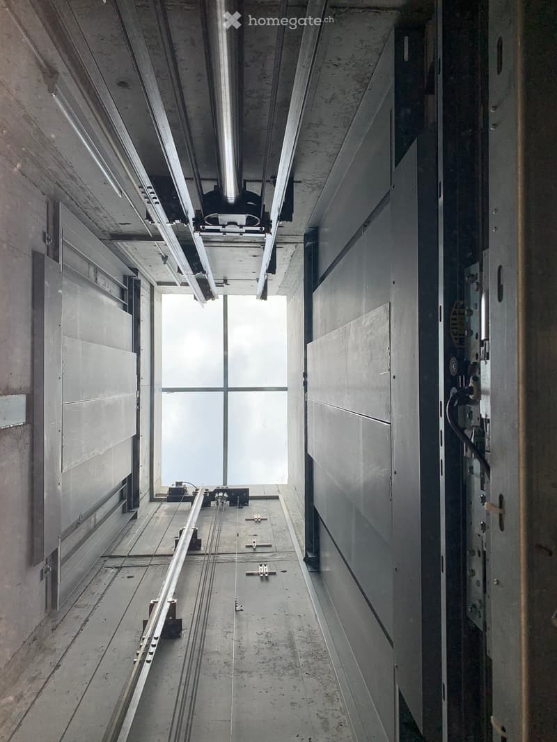A modern, minimalist corridor with concrete walls and ceiling, featuring exposed structural elements and a large window at the end allowing natural light to enter.