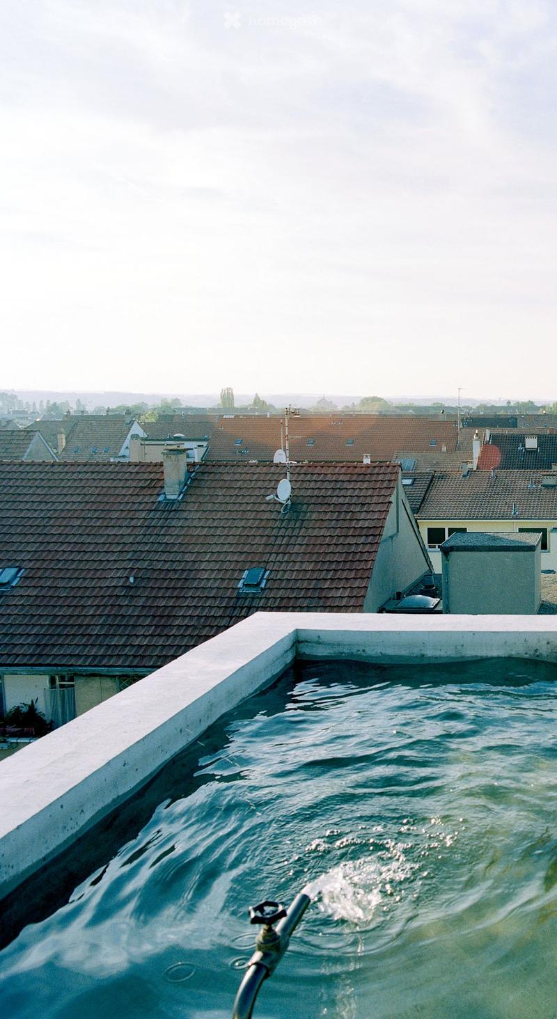 A rooftop area with a pool of water, surrounded by a low white wall, with neighboring rooftops and chimneys visible in the background.