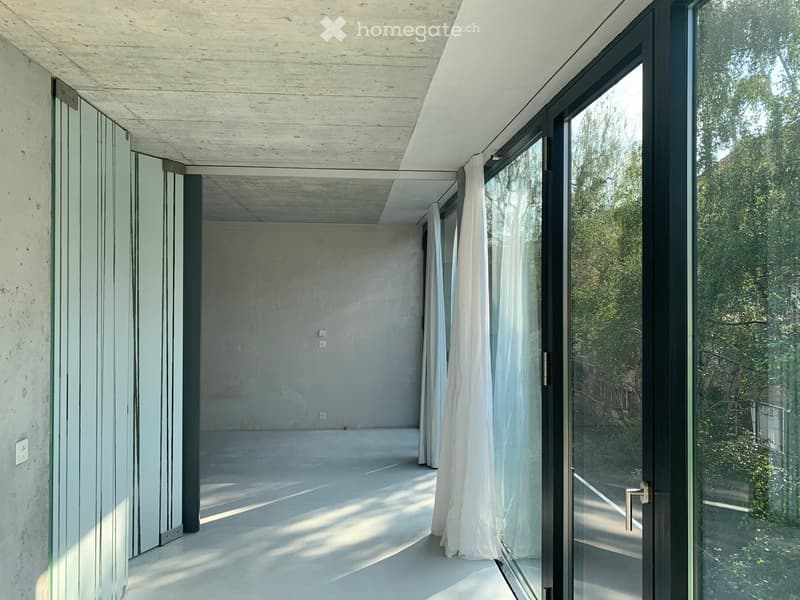 Empty room with concrete walls and floor, large glass sliding doors leading to an outdoor area, white curtains partially drawn.