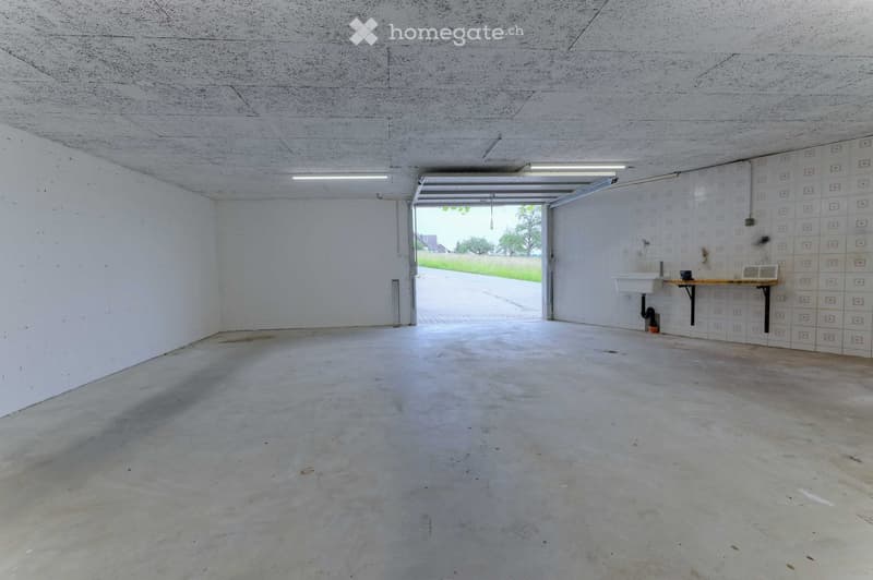 Empty garage with white walls, concrete floor, open door, small sink, and ceiling lights