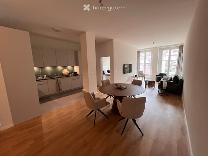 Open-plan living and dining area with light wood flooring, modern grey kitchen cabinets, a round dining table with four chairs, a black sofa, and a television mounted on the wall.