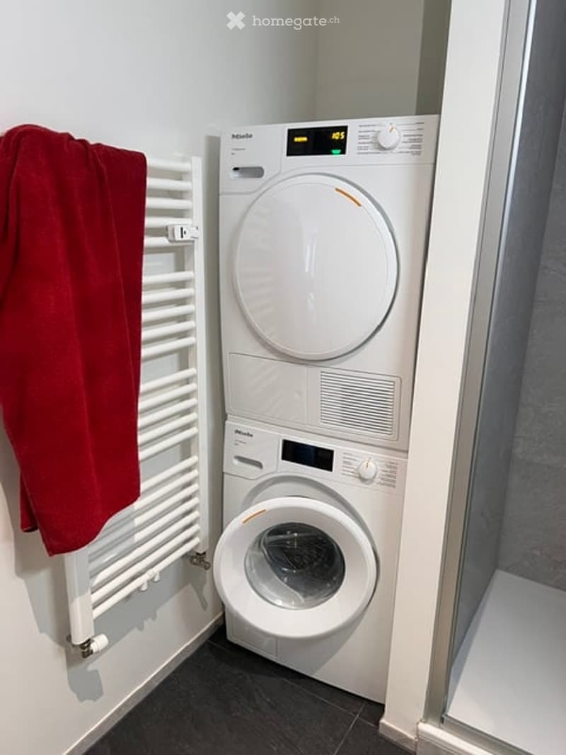Compact bathroom with a stacked washing machine and dryer unit, a white towel rack with a red towel, and a dark tiled floor.