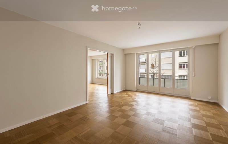 Empty room with wooden flooring, large windows, and a door leading to another space.