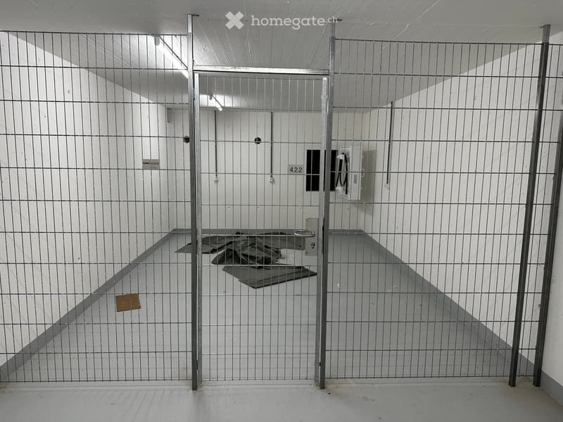 Empty room enclosed by metal bars, with a concrete floor and white walls. There is a door in the background and a small mat on the floor.
