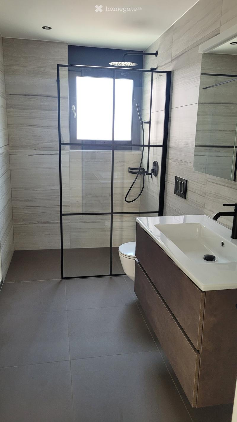 Modern bathroom with a glass-enclosed shower, a white sink with a dark faucet, and a toilet. The walls are tiled in a light, marble-like pattern, and the floor is dark grey tiles.