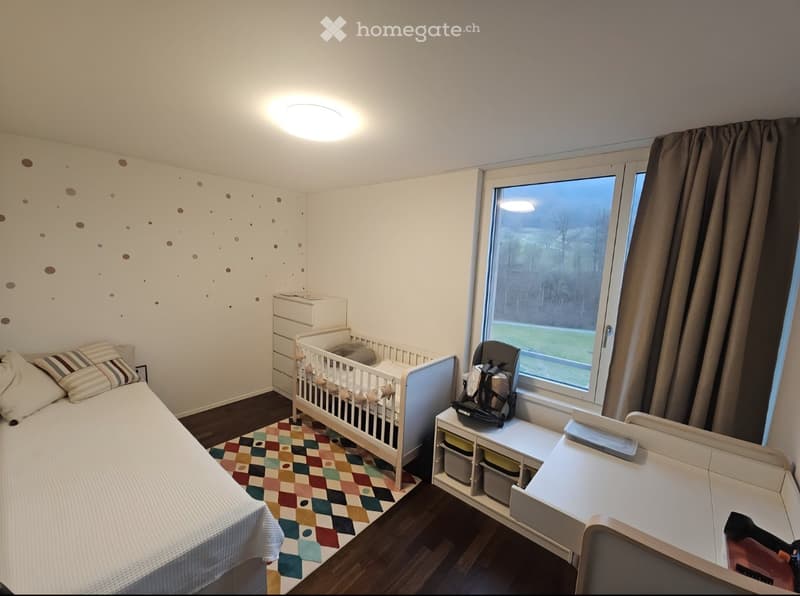 Cozy bedroom with a single bed, crib, nightstand, and a colorful rug. Features a large window with a view of the outdoors.