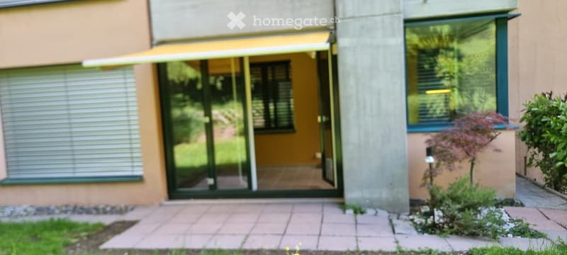 Modern building entrance with glass sliding doors, covered awning, and small garden area with plants and a tree.