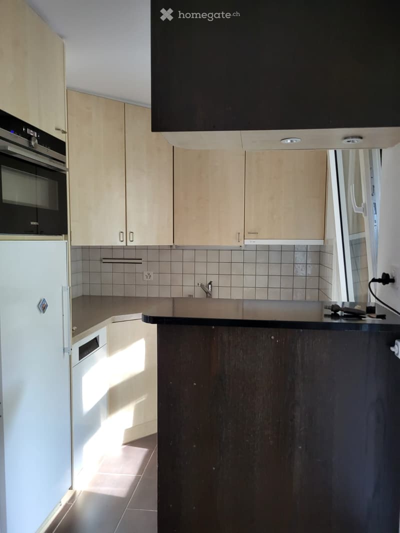 Modern kitchen with light wood cabinets, stainless steel appliances, and a dark countertop. Features include a refrigerator, oven, sink, and faucet.