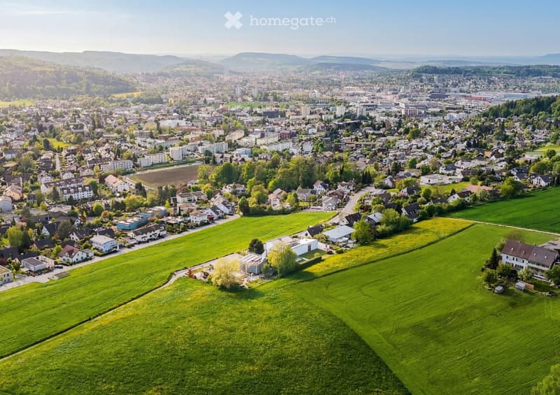 Aerial view of a suburban town with residential houses, green fields, and distant mountains under a clear sky.