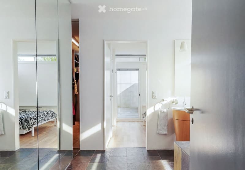 Bright, minimalist hallway with white walls, large windows, and a glass door leading to a bedroom area. Features a wooden vanity with a sink, a towel rack, and a mirror.