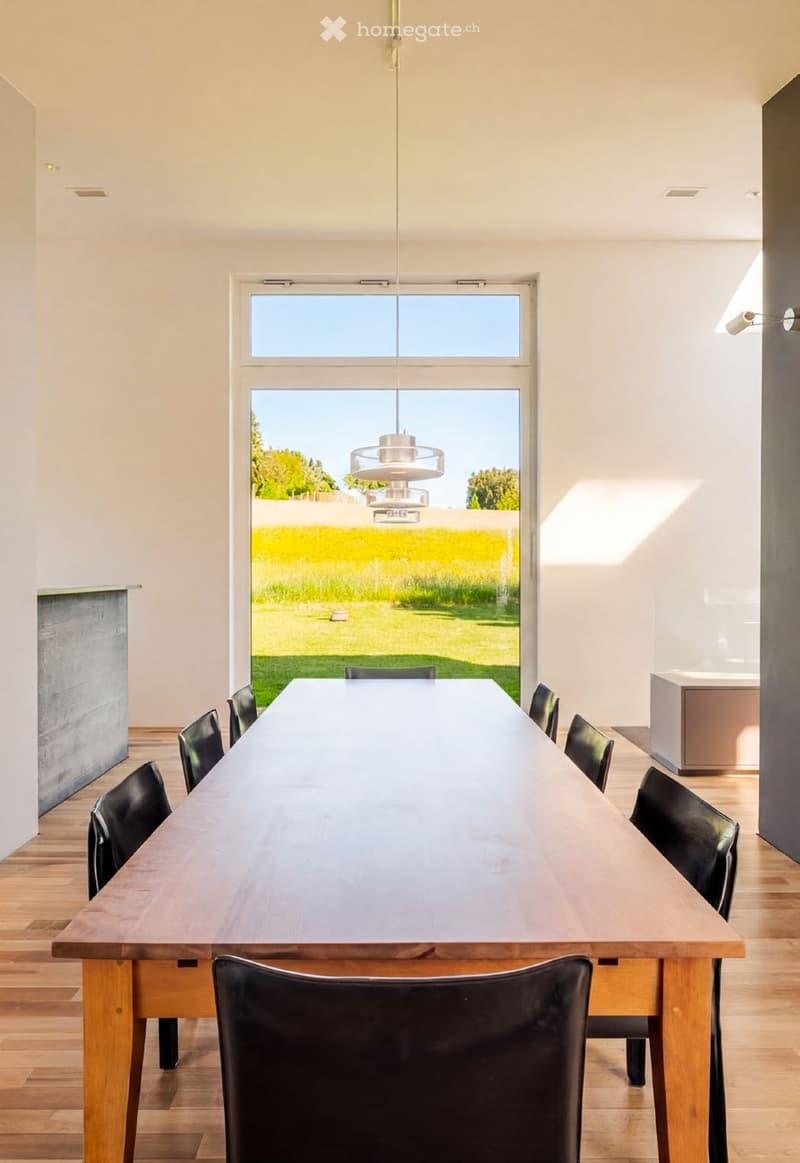 Bright dining room with a long wooden table surrounded by black chairs, pendant light fixture, large window overlooking a green field.