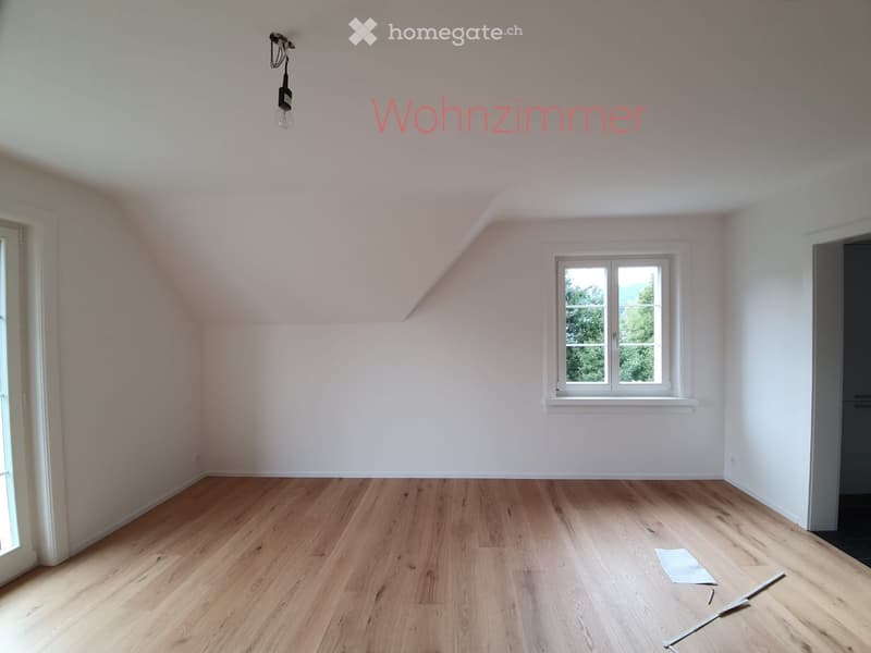 Empty living room with wooden flooring, white walls, and a single window showing greenery outside. No furniture or equipment present.